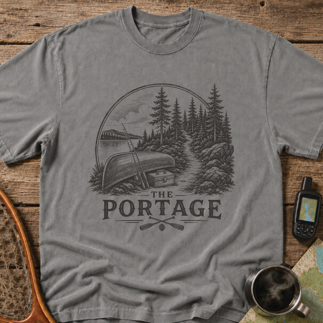 The Portage t-shirt featuring an overturned canoe beside a forest trail with fishing gear.