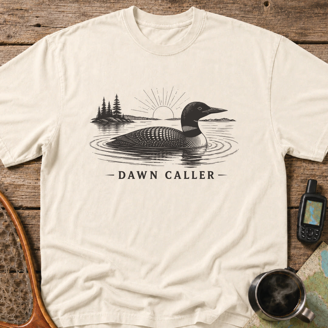 Dawn Caller loon t-shirt featuring a detailed loon floating on a northern lake at sunrise with wilderness shoreline artwork.